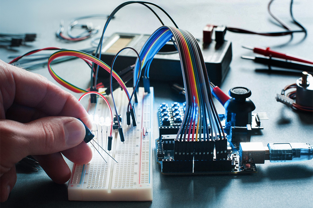 Close-up of a hand placing an electronic component into a breadboard connected to a microcontroller board, with multicolored jumper wires and illuminated LEDs on a workbench surrounded by small tools.