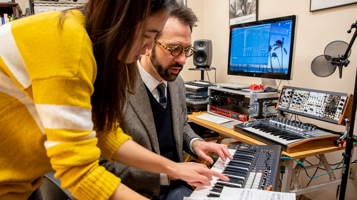 A student and a professor work together over a keyboard with various music technology equipment visible in the background.