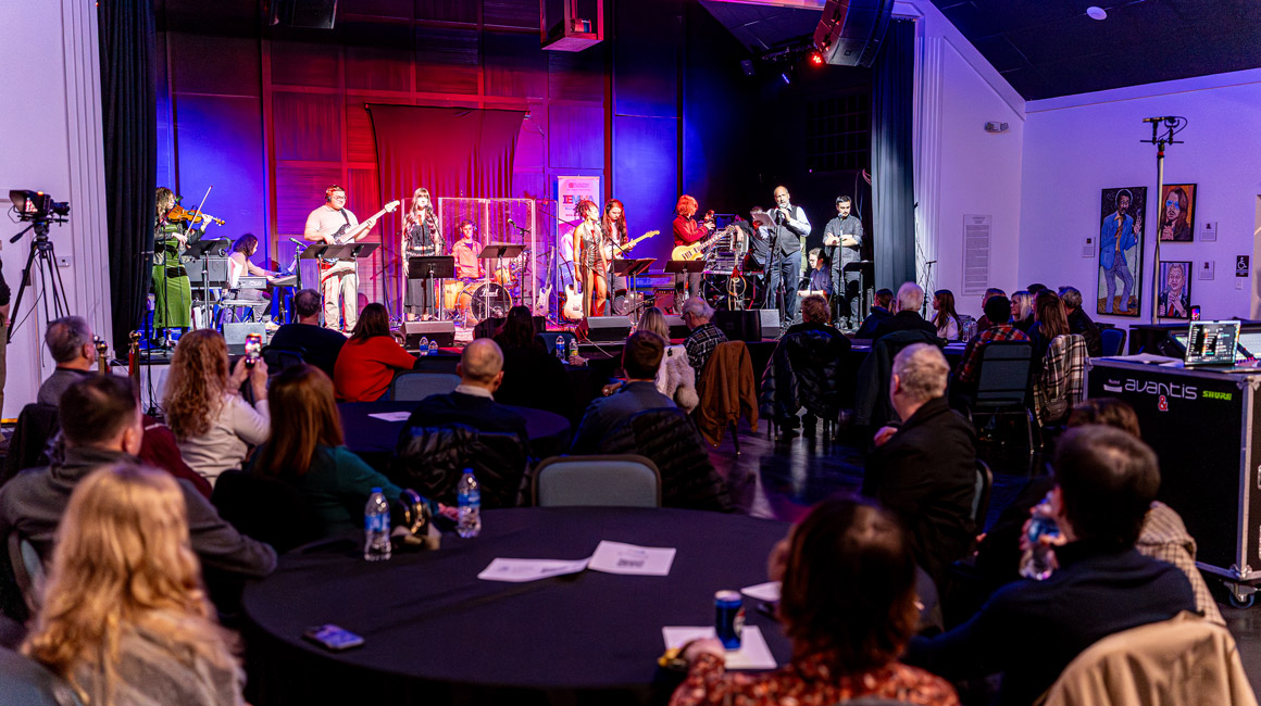 An audience is seen from behind watching a concert happening on a stage.