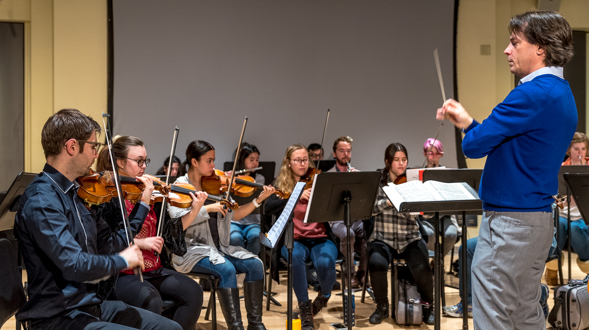 Man in a blue sweater conducts a chamber orchestra.