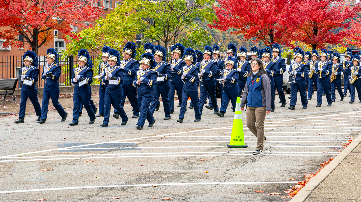 A marching band marches in front of colorful autumn trees.