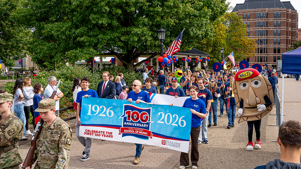 Dean David Allen Wehr and two music student ambassadors lead the Duquesne Pep Band with a Centennial Celebration banner. They march in a parade on Academic Walk.