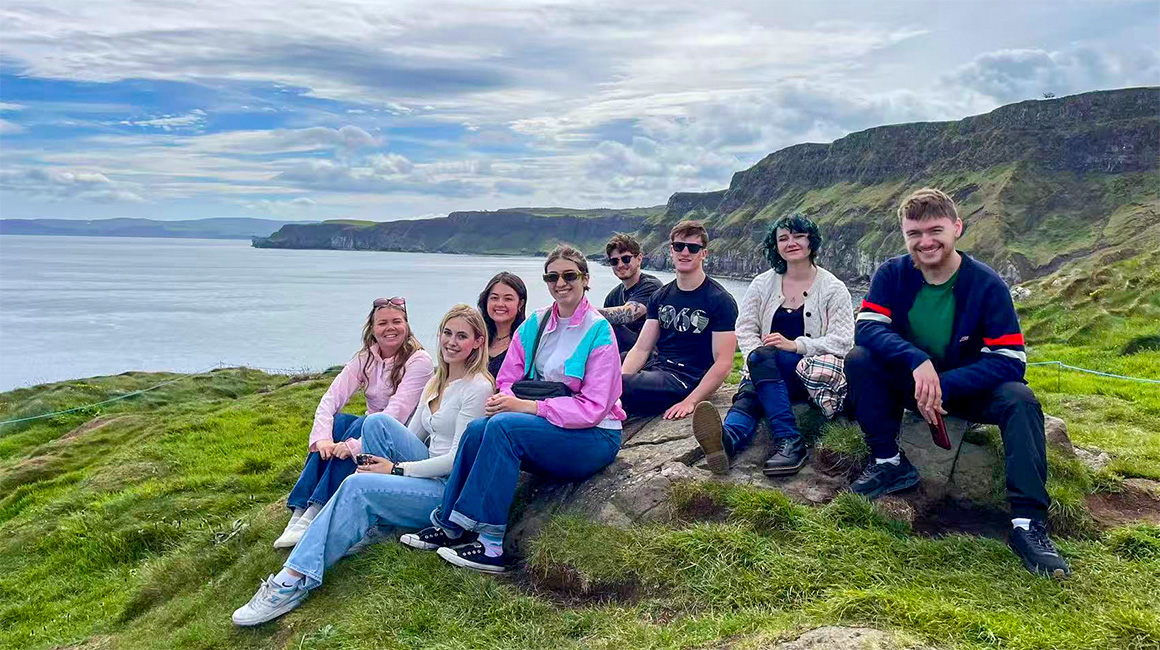 A group of eight young adults sits together on a grassy rock overlooking a dramatic coastal landscape of steep green cliffs and calm blue water. The group smiles toward the camera while layered clouds stretch across the sky, creating soft, diffused light. The rugged shoreline curves into the distance, emphasizing the scenic overlook and expansive ocean view.