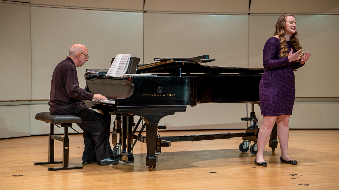 A woman in a purple dress sings in recital with a male accompanist at the piano.
