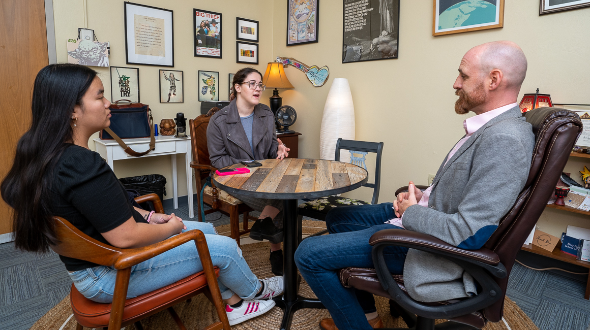 A teacher and two female students sit around a table in an office and talk.