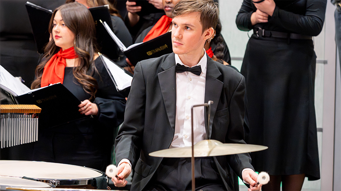 A percussionist performs in a tuxedo. A choir can be seen in the background wearing black with red scarves.