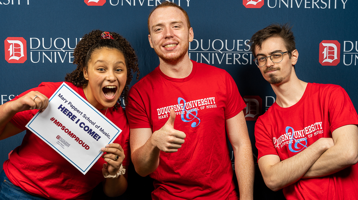 Three students pose for a photo wearing red shirts. One holds a sign that reads Mary Pappert School of Music, here I come!