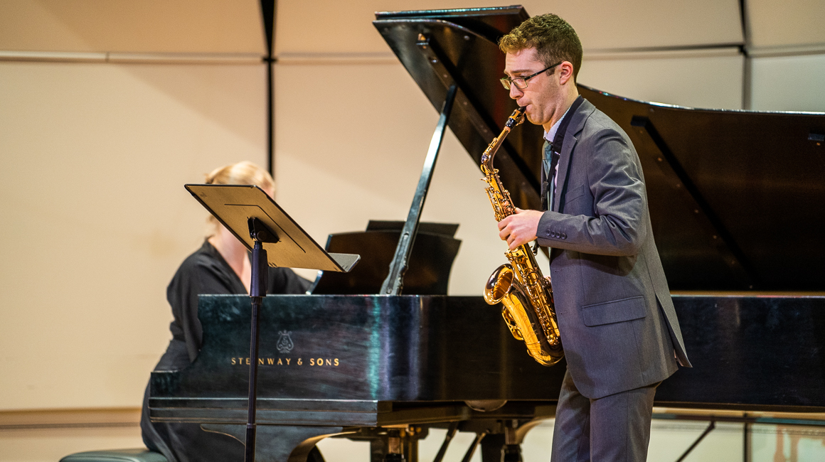 Student plays a saxophone with a pianist in the background.