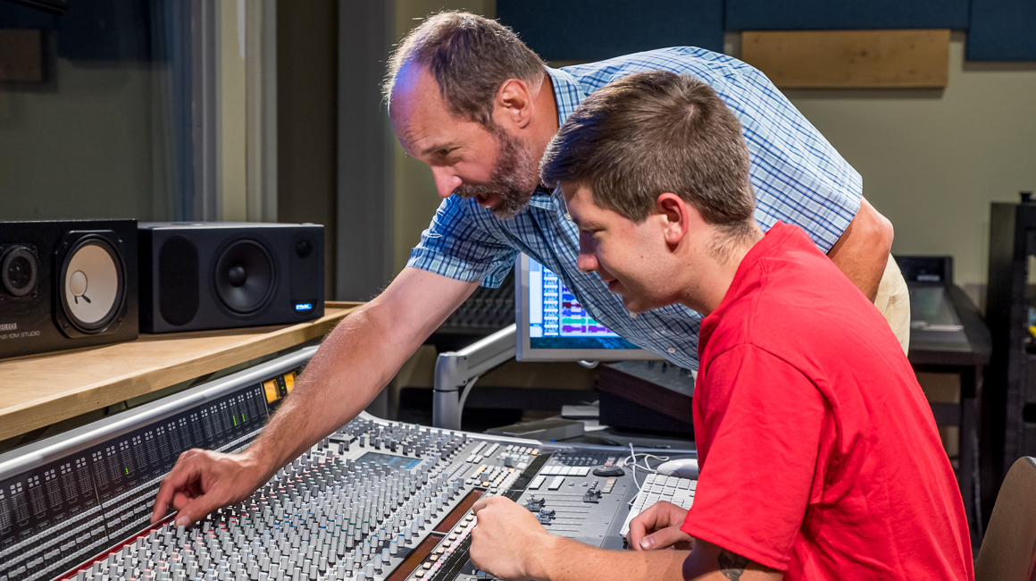 A professor shows a student a technique on a mixing console.