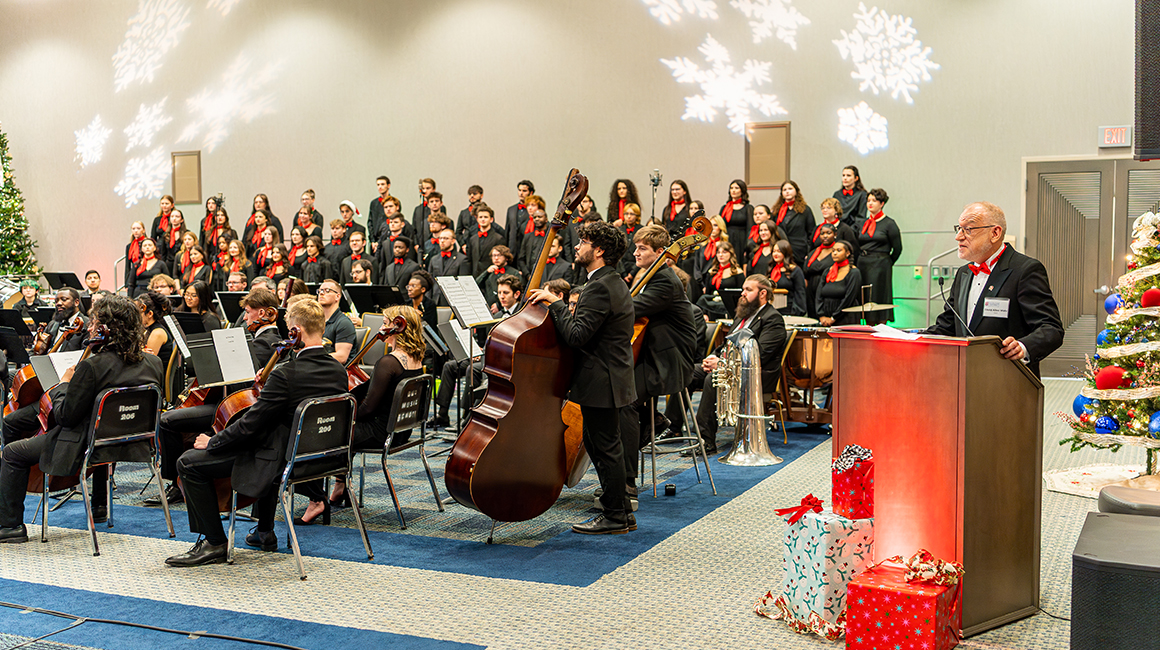 Speaker at podium addresses audience beside orchestra and choir during holiday concert.