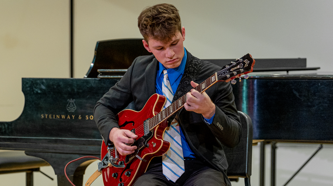 A person wearing a suit plays a red electric guitar on the stage of the PNC Recital Hall.
