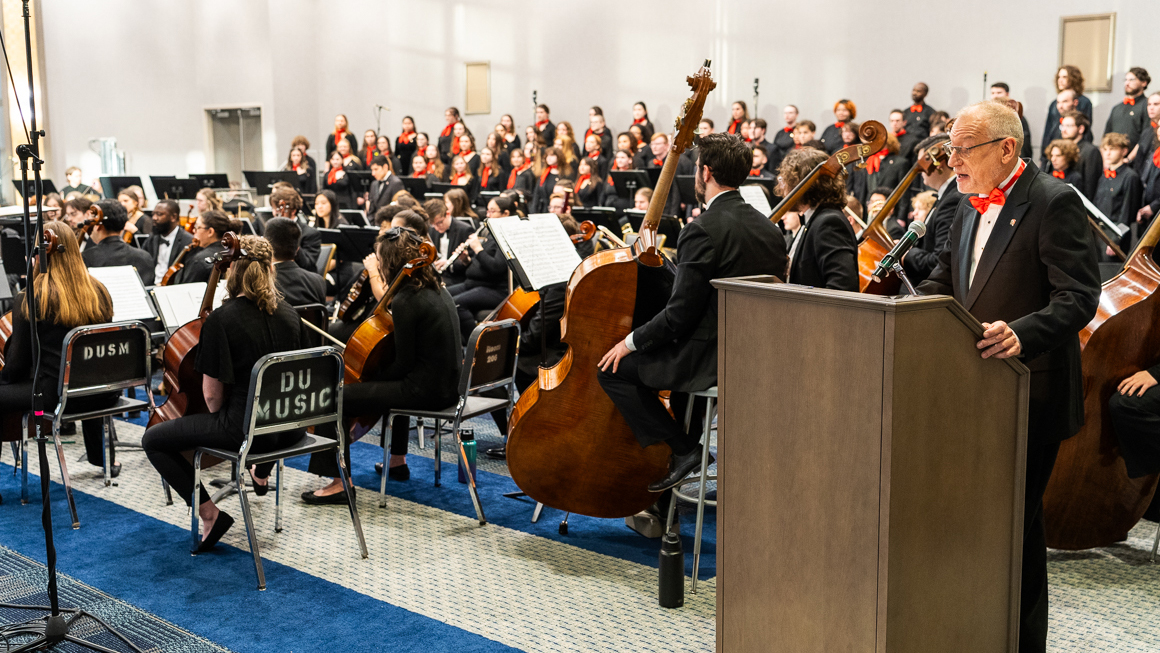 A person speaks at a podium with an ensemble in the background.