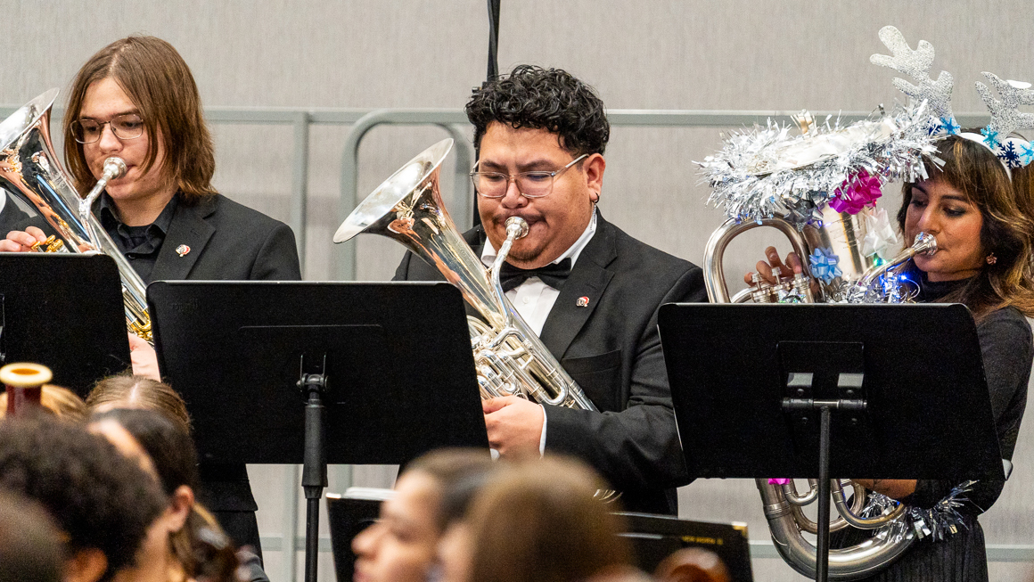 Three people play low brass instruments, one of which is decorated with tinsel.