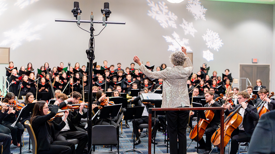 Conductor leading full orchestra and choir beneath projected snowflakes during a Christmas performance.
