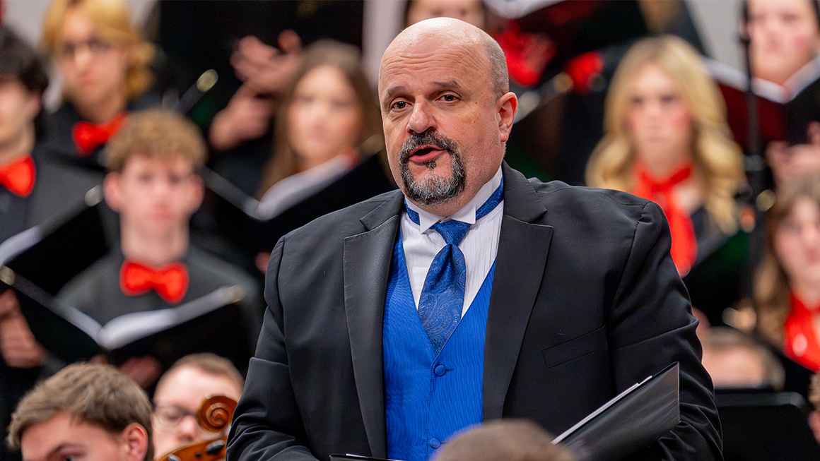 Male soloist in blue vest singing in front of choir during holiday concert.