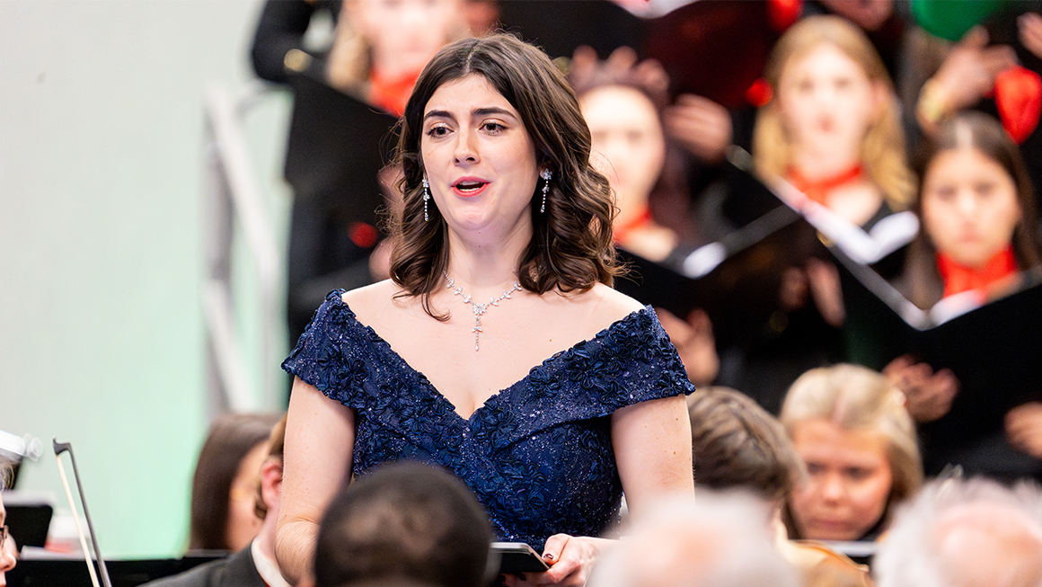 Female soloist in navy gown singing with orchestra and choir behind her.