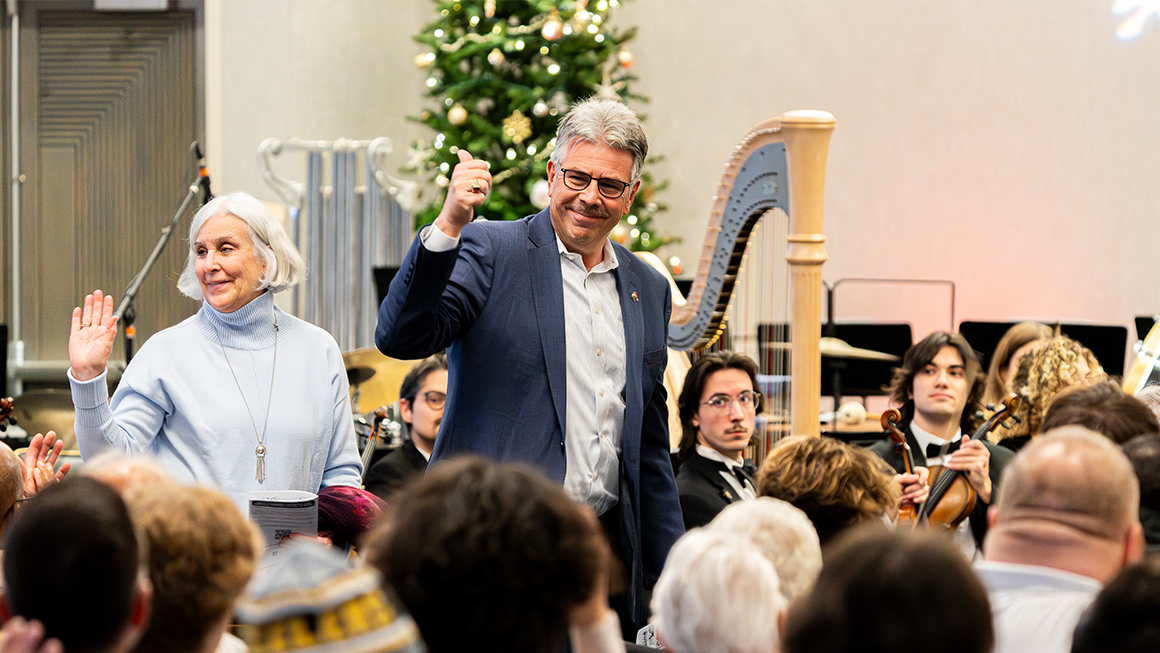 Two guests acknowledge applause with raised hands in front of orchestra and harp.