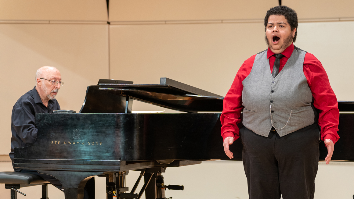 A student in a red shirt and gray vest sings with a pianist.