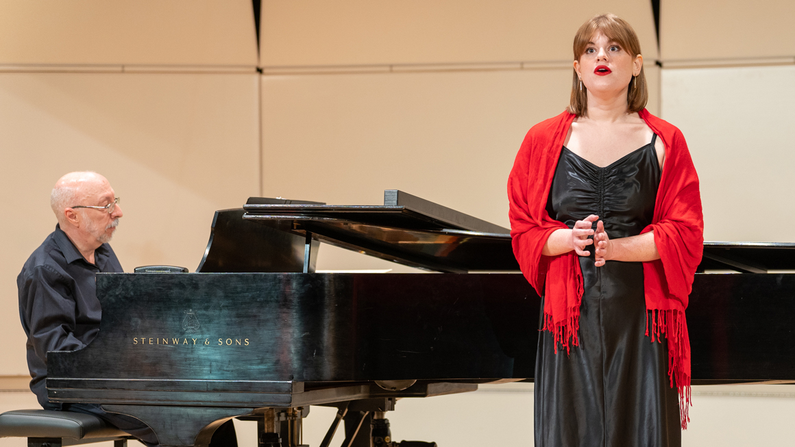 A woman in a black dress with red accent sings on stage with a pianist.