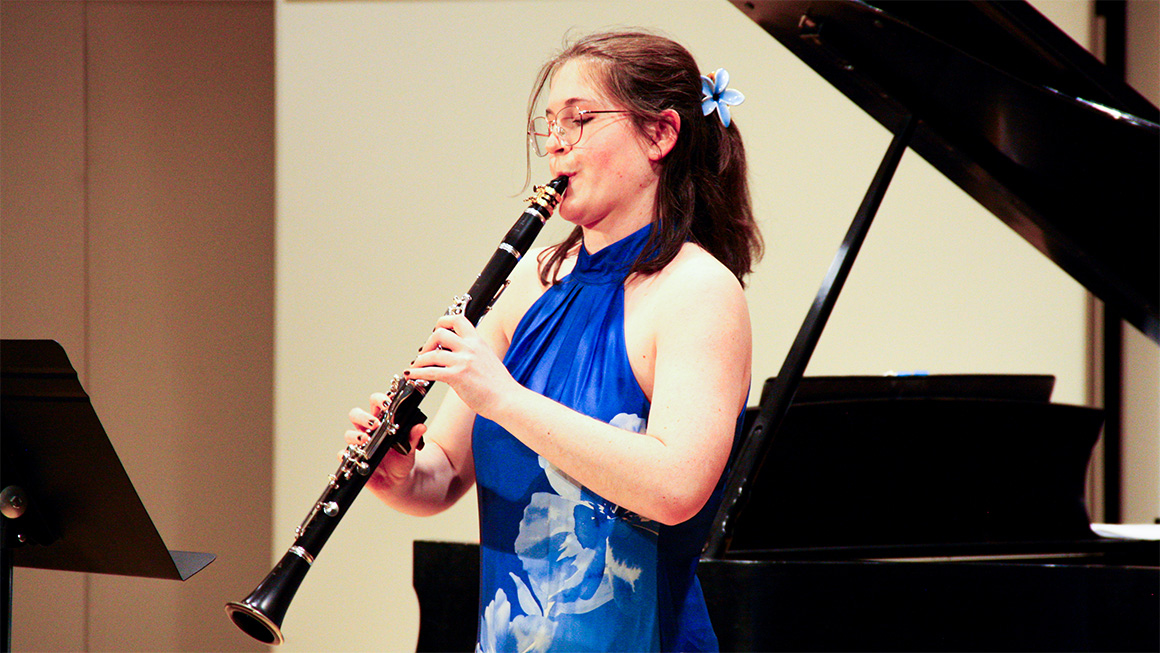 A person with glasses and long hair wearing a blue dress plays clarinet.