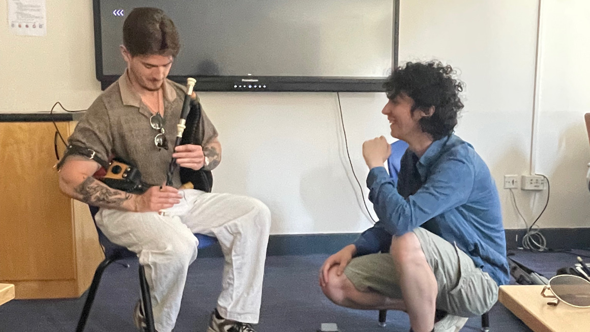 Two people share a conversation during a traditional Irish music session, with one seated and demonstrating a set of uilleann pipes while the other crouches nearby listening and smiling.