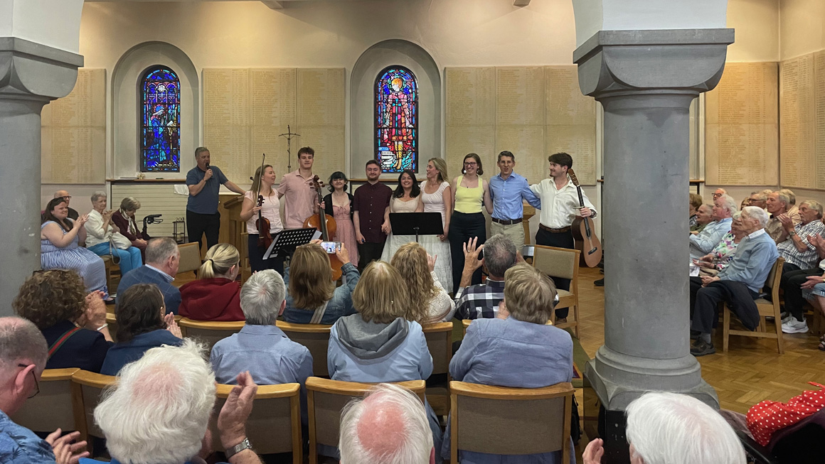 Student musicians and faculty stand before an applauding audience following a performance inside a church. Stained glass windows frame the ensemble as audience members applaud from their seats.