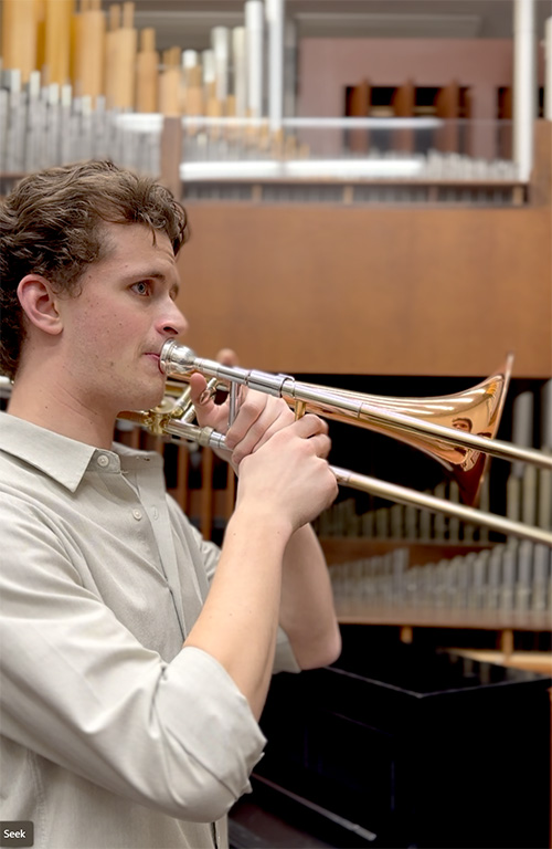 A person plays a trombone in front of a pipe organ.