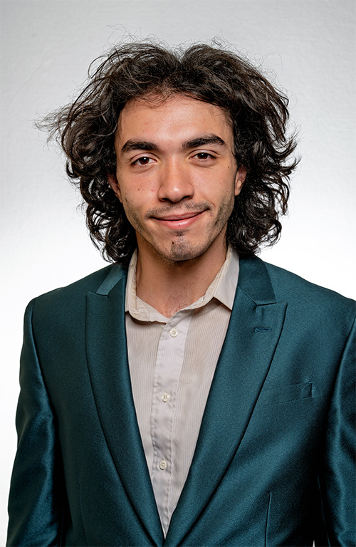 A person wearing a teal blazer and white collared shirt smiles for a headshot in front of a neutral background.