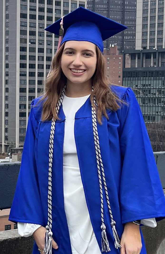 Student in graduation cap and gown stands in front of a city background.