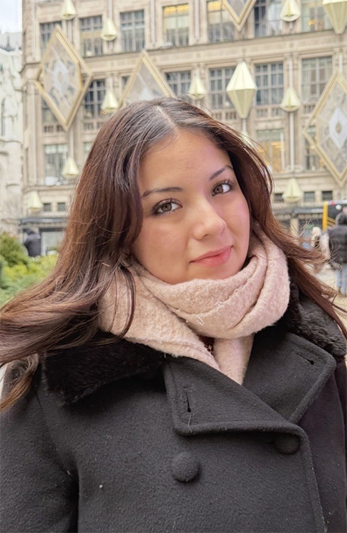 A person wears a black wool coat and pink scarf while posing for a photo in front of a building.