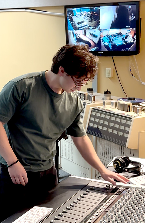 A person manipulates a recording console. Camera feeds from adjacent rooms are shown on a TV in the background.