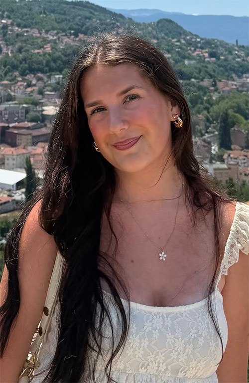 A person with long dark hair wearing a white top smiles for a picture in front of a picturesque background.
