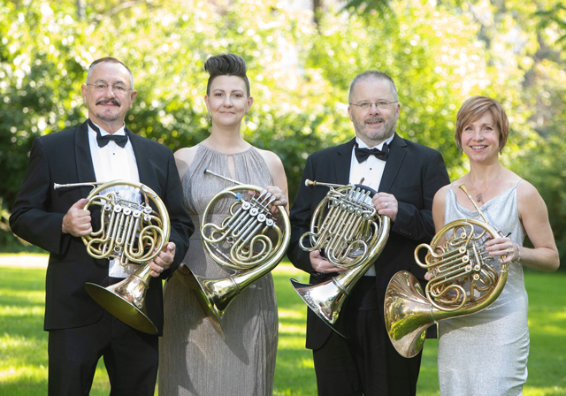 Four people hold French horns for an outdoor portrait.
