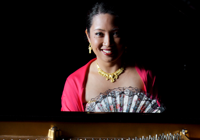 A person wearing an ornate necklace and a pink top poses at a piano.