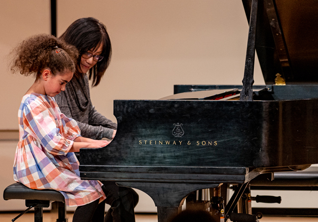 A child plays a piano next to their teacher.