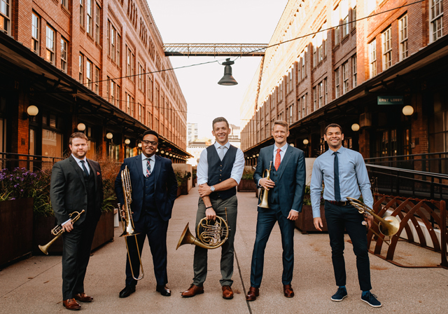 Five people with brass instruments pose in an industrial location.