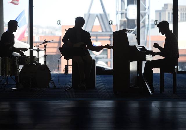Silhouettes of three musicians—a drummer, guitarist, and pianist—can be seen in front of a glass wall overlooking Duquesne's campus.