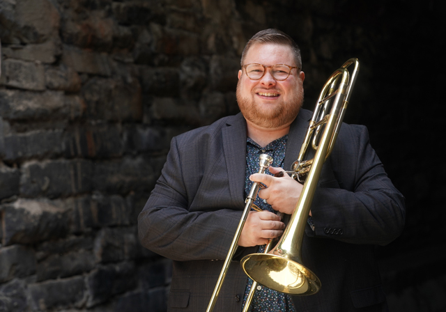 A person with a trombone poses in front of a brick wall.