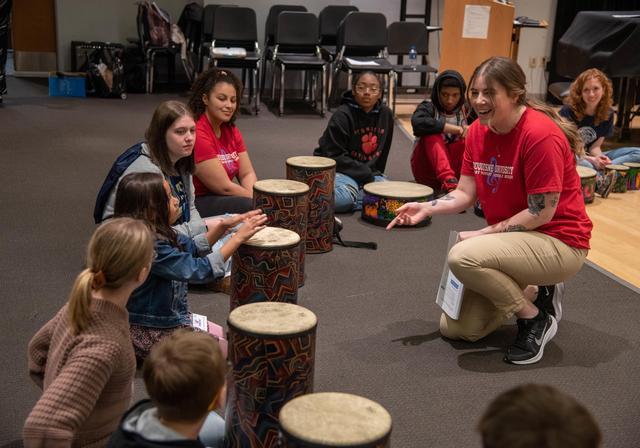 A smiling person leads a drum circle with several young students.