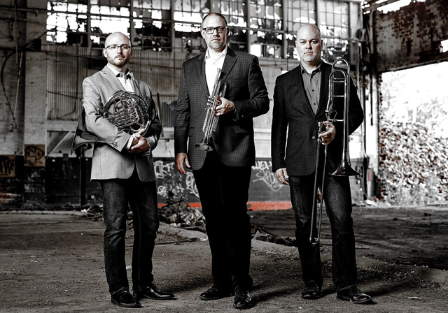 A trio of people hold brass instruments in a warehouse.