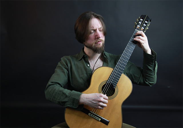 A person plays a classical guitar in front of a neutral background.