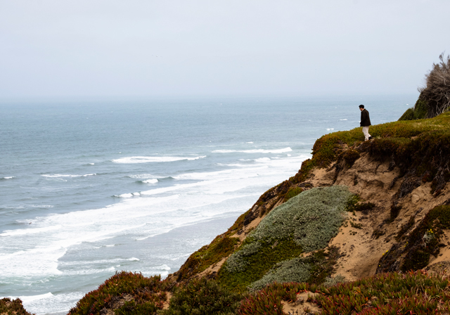 A lone figure walks along a rugged coastal cliff, overlooking rolling ocean waves beneath a pale, overcast sky, with textured sand and low vegetation framing the shoreline.