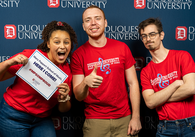 Three students in red MPSOM shirts post for a photo, one holds a sign that reads Mary Pappert School of Music, here I come!