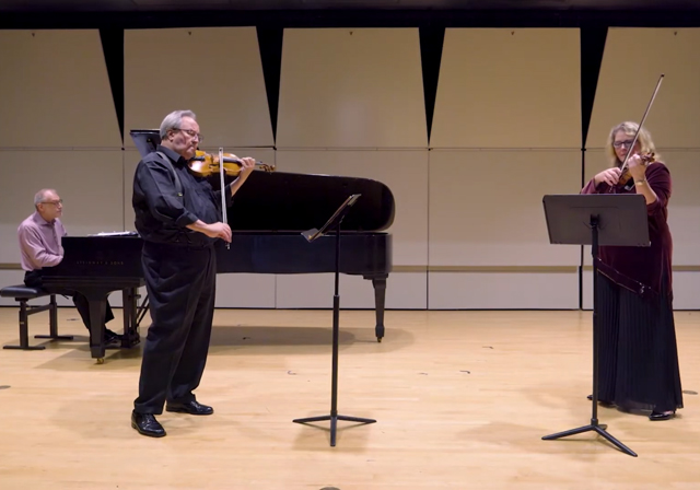 Two violinists and a pianist perform on a stage.
