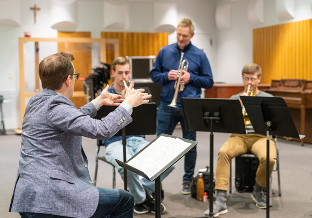 A man conducts two trumpet players while another man looks on from behind.