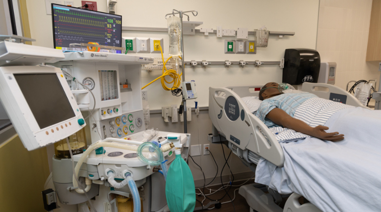 An operating room machine next to a hospital bed with a mannequin laying on it.