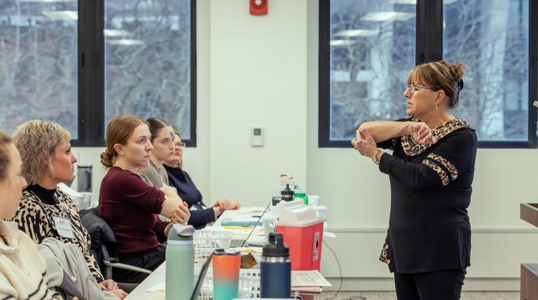 A woman in front of a classroom of students. She is pointing at her elbow. The students are watching intently.