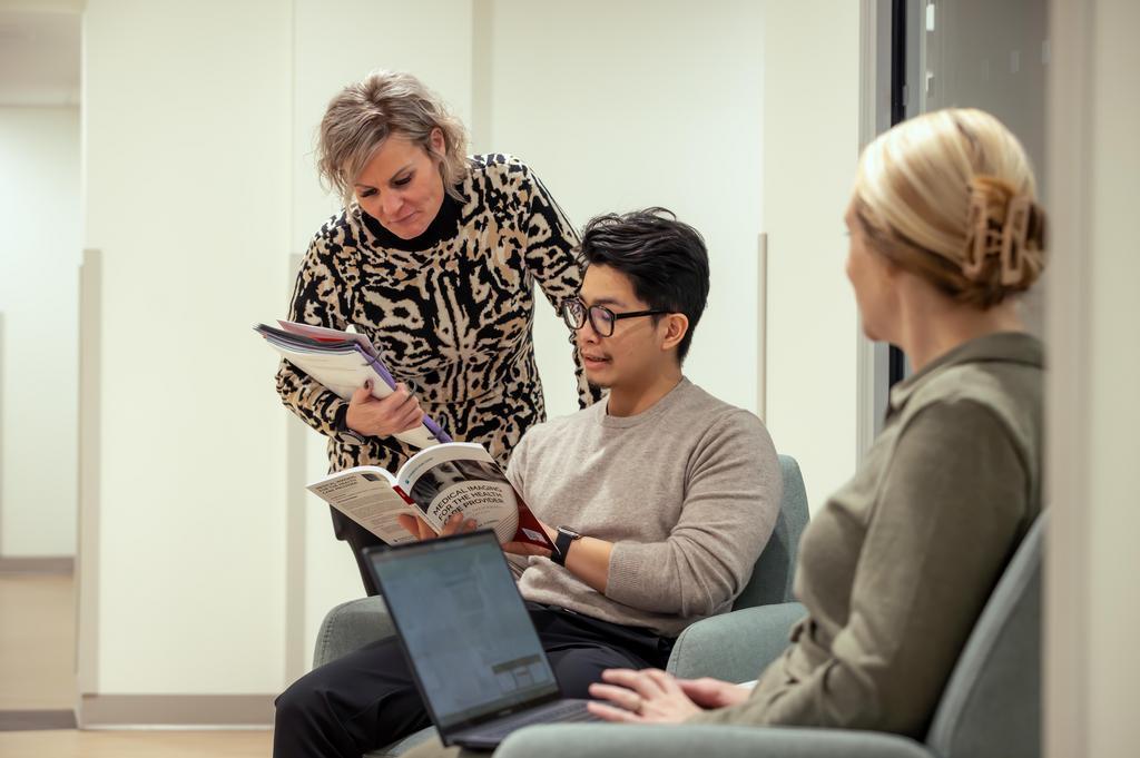 Two people sitting in chairs, one on a laptop and one reading a book. The person reading the book is pointing at the book to someone standing over them.