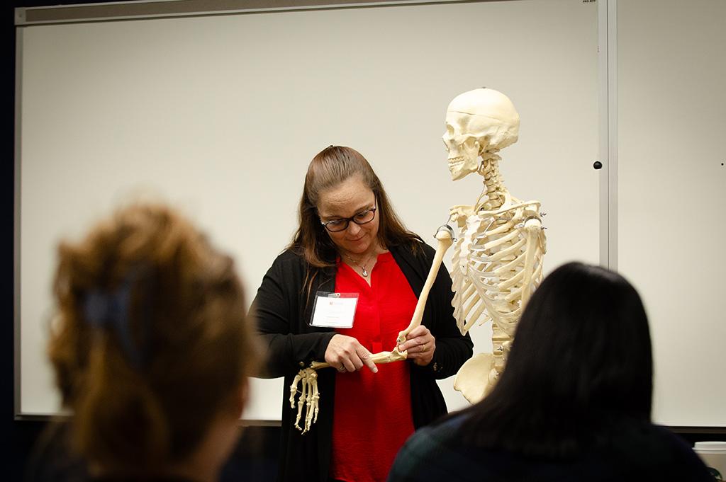 A woman in front of a room pointing at a classroom skeleton.