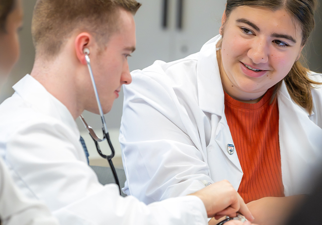 student in lab coat examining another student with a stethoscope